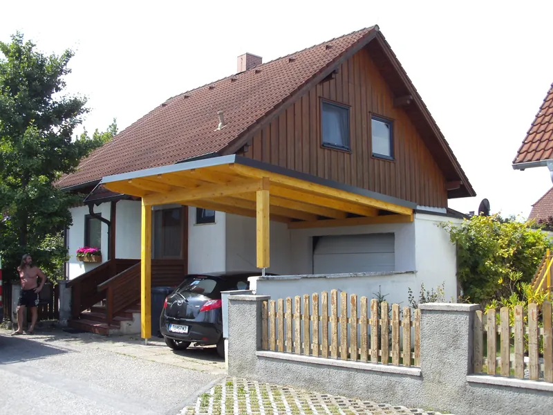 Carport mit Holzdach und gelb lasierten Stützen vor einem Einfamilienhaus